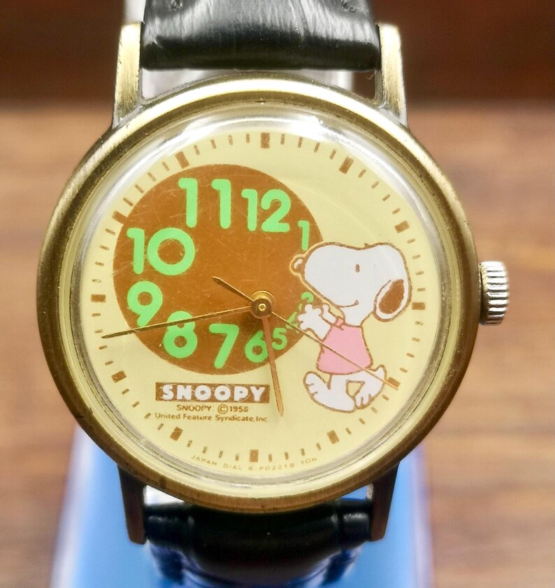 May include: A wristwatch with a round brass-toned case and a black leather strap. The cream-colored dial features a brown circle with large green numbers, a graphic of Snoopy wearing a pink shirt, and the text SNOOPY 1958 United Feature Syndicate, Inc.