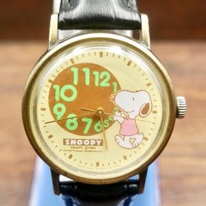 Puede incluir: Un reloj de pulsera analógico redondo con caja de metal dorado y correa de cuero negro. La esfera color crema presenta un círculo marrón, números verdes y un gráfico de Snoopy con una camisa rosa. El texto incluye SNOOPY y United Feature Syndicate Inc.
