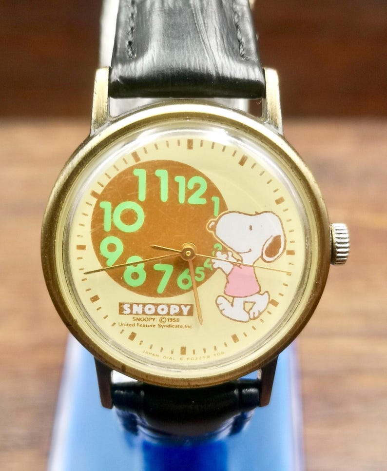May include: A round analog wristwatch with a gold-tone metal case and a black leather strap. The cream-colored dial features a brown circle, green numerals, and a graphic of Snoopy wearing a pink shirt. Text includes SNOOPY and United Feature Syndicate Inc.