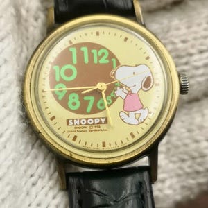 May include: A wristwatch with a black leather strap and a gold-tone metal case. The watch face features a cream background, a brown center circle, and large green numbers. Snoopy is depicted wearing a pink shirt. Text includes SNOOPY and United Feature Syndicate, Inc.
