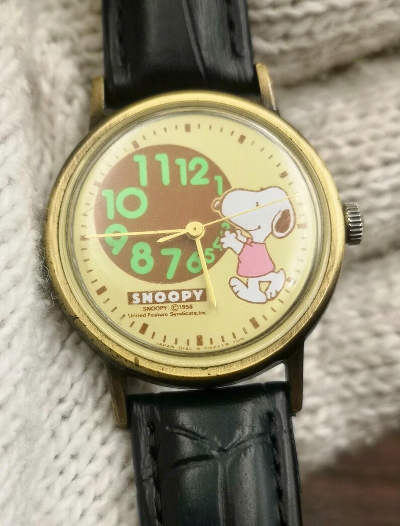 May include: A wristwatch with a black leather strap and a gold-tone metal case. The watch face features a cream background, a brown center circle, and large green numbers. Snoopy is depicted wearing a pink shirt. Text includes SNOOPY and United Feature Syndicate, Inc.