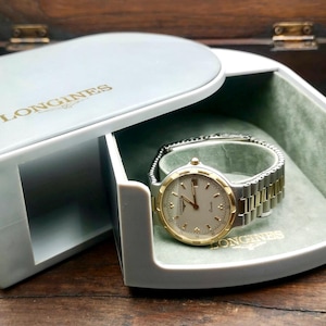 May include: A Longines watch with a gold-tone bezel and silver-tone band rests in its open, light gray box. The watch has a white face with gold hands and markers. The box is lined with green velvet.