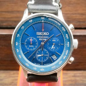 Puede incluir: Un reloj cronógrafo Seiko de acero inoxidable con esfera azul y correa de cuero negro. La esfera presenta múltiples subesferas, una escala taquimétrica y el texto "SEIKO CHRONOGRAPH 100M". La caja es plateada.