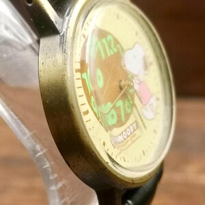 May include: An analog wristwatch with a brass-toned metal case and a black leather strap. The watch face features a cream background with a graphic of Snoopy, green numerals, and the text "SNOOPY" printed in a white bone-shaped graphic at the bottom.