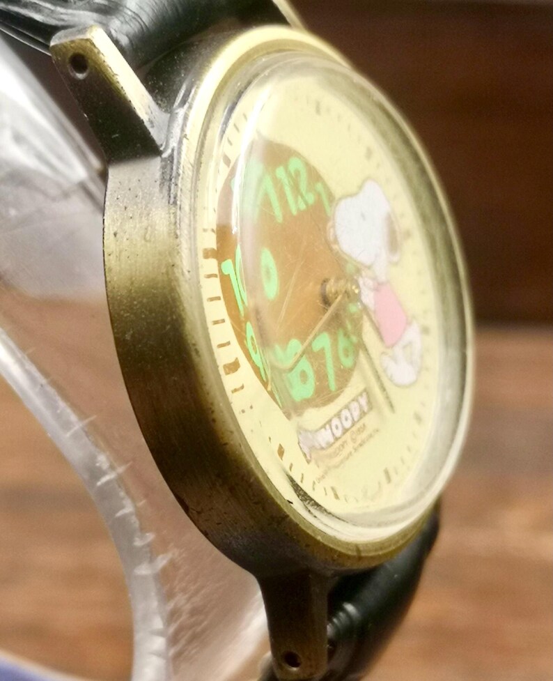 May include: An analog wristwatch with a brass-toned metal case and a black leather strap. The watch face features a cream background with a graphic of Snoopy, green numerals, and the text "SNOOPY" printed in a white bone-shaped graphic at the bottom.