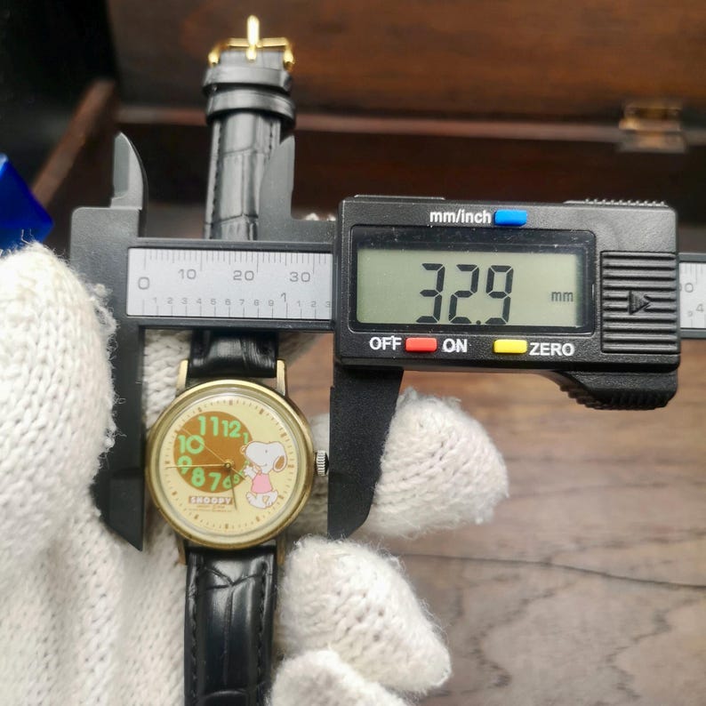 May include: A round analog wristwatch with a black leather strap and a cream-colored face featuring a Snoopy graphic and large green numerals. A black digital caliper is measuring the watch case, displaying a width of 32.9 mm on its screen.