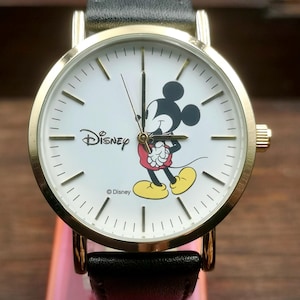 Puede incluir: Un reloj de pulsera dorado con una esfera blanca con Mickey Mouse. El reloj tiene una correa de cuero negro y el logotipo de Disney. Las manecillas del reloj son negras y los marcadores de hora son líneas negras.