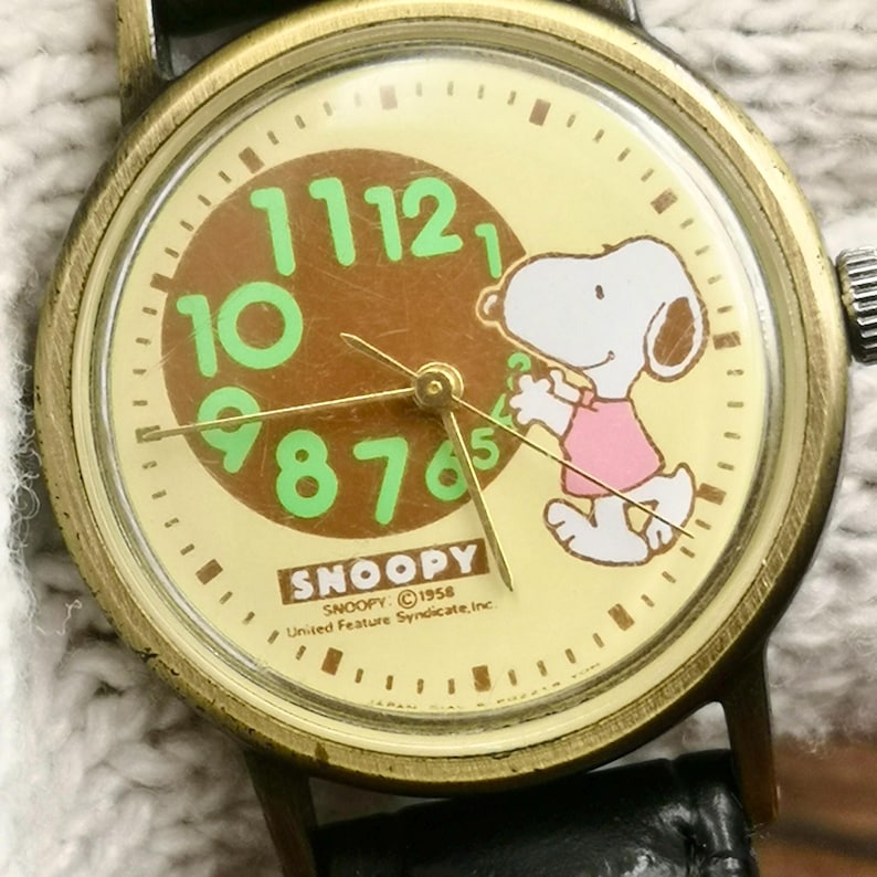 May include: A vintage-style analog wristwatch with a brass-toned metal case and a black leather strap. The cream-colored dial features a graphic of Snoopy, light green numerals, gold-tone hands, and the text SNOOPY &copy; 1958 United Feature Syndicate, Inc.