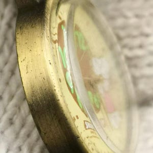 May include: A round pocket watch with a distressed bronze-toned metal casing and a domed glass crystal. The watch face features a cream-colored background with light green numerals and a small graphic illustration of a white figure with pink accents.