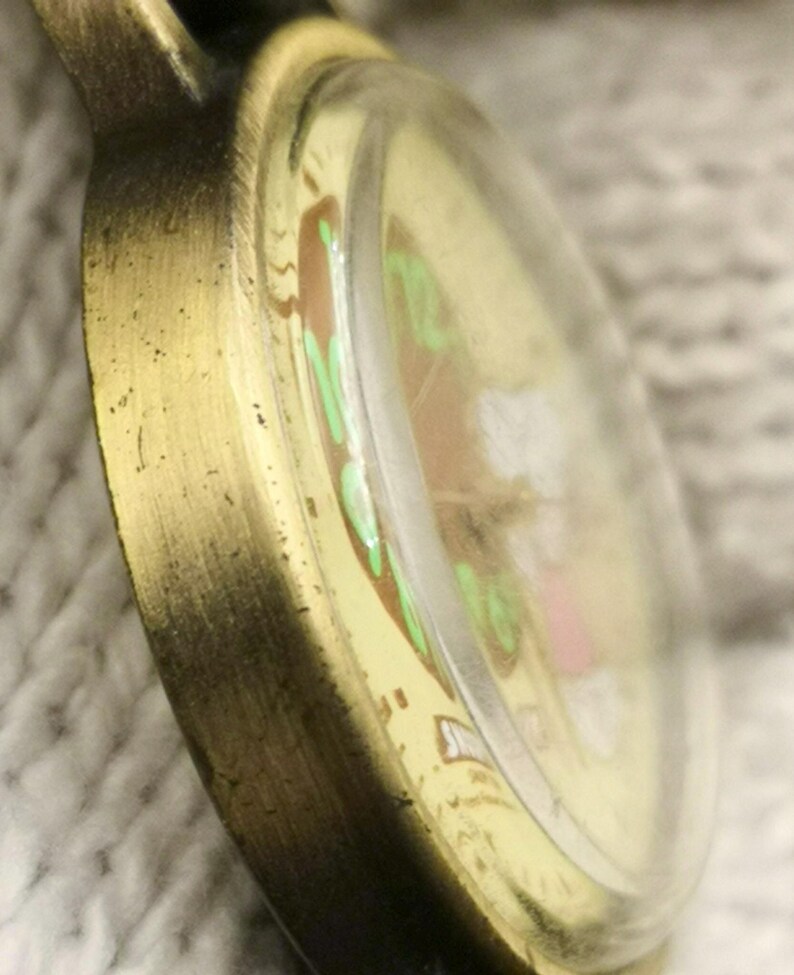 May include: A round pocket watch with a distressed bronze-toned metal casing and a domed glass crystal. The watch face features a cream-colored background with light green numerals and a small graphic illustration of a white figure with pink accents.