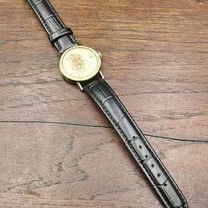May include: A wristwatch with a gold-tone circular case and a black leather strap featuring a textured crocodile pattern. The watch face displays a light-colored background with dark numerals and a small graphic illustration of a character.