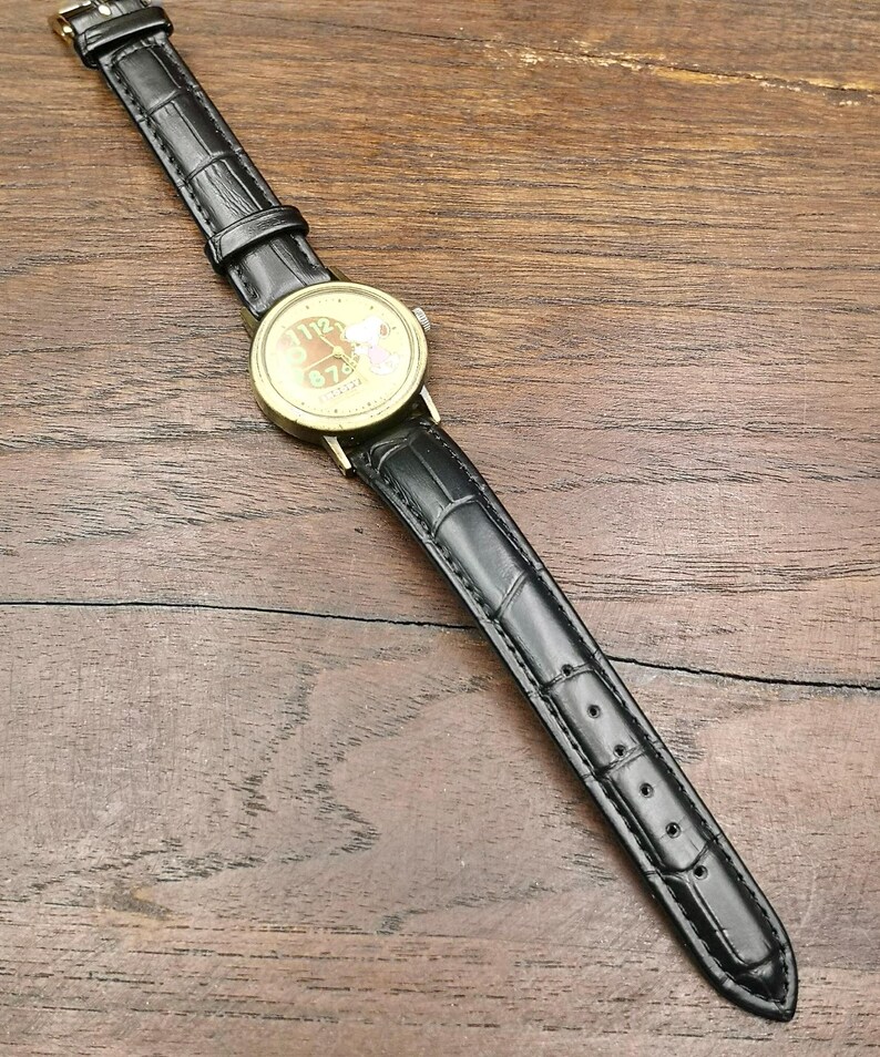 May include: A wristwatch with a gold-tone circular case and a black leather strap featuring a textured crocodile pattern. The watch face displays a light-colored background with dark numerals and a small graphic illustration of a character.