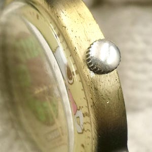 May include: A gold-toned metal wristwatch case with a textured finish and a small, round, silver-colored crown on the side. The watch face features a light-colored background with visible green and pink graphic elements under the glass crystal.