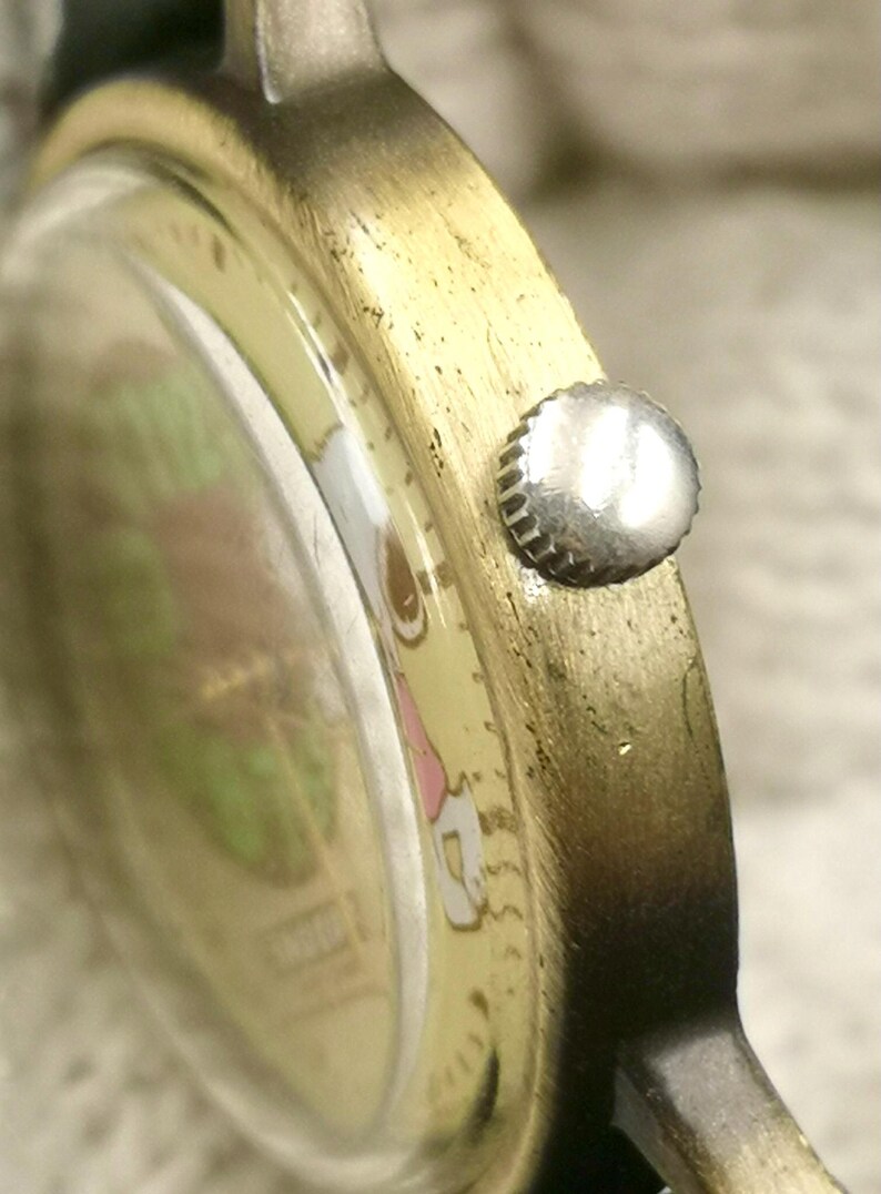 May include: A gold-toned metal wristwatch case with a textured finish and a small, round, silver-colored crown on the side. The watch face features a light-colored background with visible green and pink graphic elements under the glass crystal.