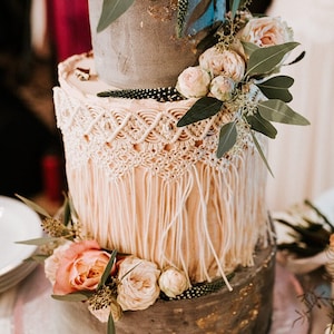May include: A three-tiered wedding cake with a rustic, bohemian theme. The cake is decorated with a macrame-style fringe, blush pink roses, and greenery. The bottom tier is a gray concrete-like finish with gold accents.