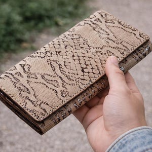 May include: A beige wallet with a snakeskin pattern. The rectangular wallet has a flap closure. The edges are a darker brown, and the wallet is held in a hand.