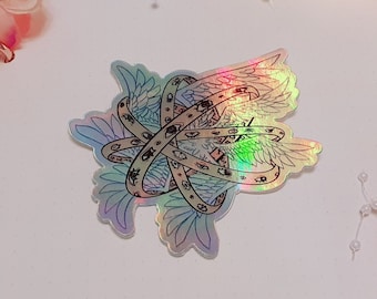 May include: A holographic sticker featuring an intricate design of multiple rings and wings, with eyes incorporated throughout. The sticker is placed on a white surface, surrounded by pink floral decorations and fairy lights.