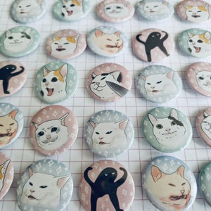 May include: A selection of round pin badges displaying cat meme illustrations. The badges depict diverse cat expressions and poses, set against pastel-coloured backgrounds with diamond patterns. Some badges feature a black silhouette.