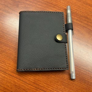 Könnte beinhalten: Ein schwarzes Ledernotizbuch mit braunem, gestepptem Rand und einem goldenen Knopfverschluss. Ein silberner Stift ist am Notizbuch befestigt.
