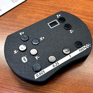 May include: A black handheld remote control featuring multiple circular buttons labeled with X+, X-, Y+, Y-, Z+, and Z- axes. The device includes a Bluetooth icon, a small rectangular display, and a white strip with speed settings marked 0.025, 0.25, 1, and FAST.