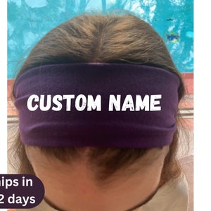 May include: A deep purple headband with the text "CUSTOM NAME" in white block letters. The headband is worn across the forehead. The background is a blurred mix of blue and green.
