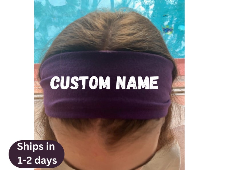May include: A deep purple headband with the text "CUSTOM NAME" in white block letters. The headband is worn across the forehead. The background is a blurred mix of blue and green.