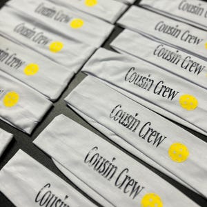 May include: A collection of light gray headbands with the text "Cousin Crew" in black script. Each headband features a yellow pickleball design. The headbands are neatly arranged, suggesting they are ready for sale or distribution.