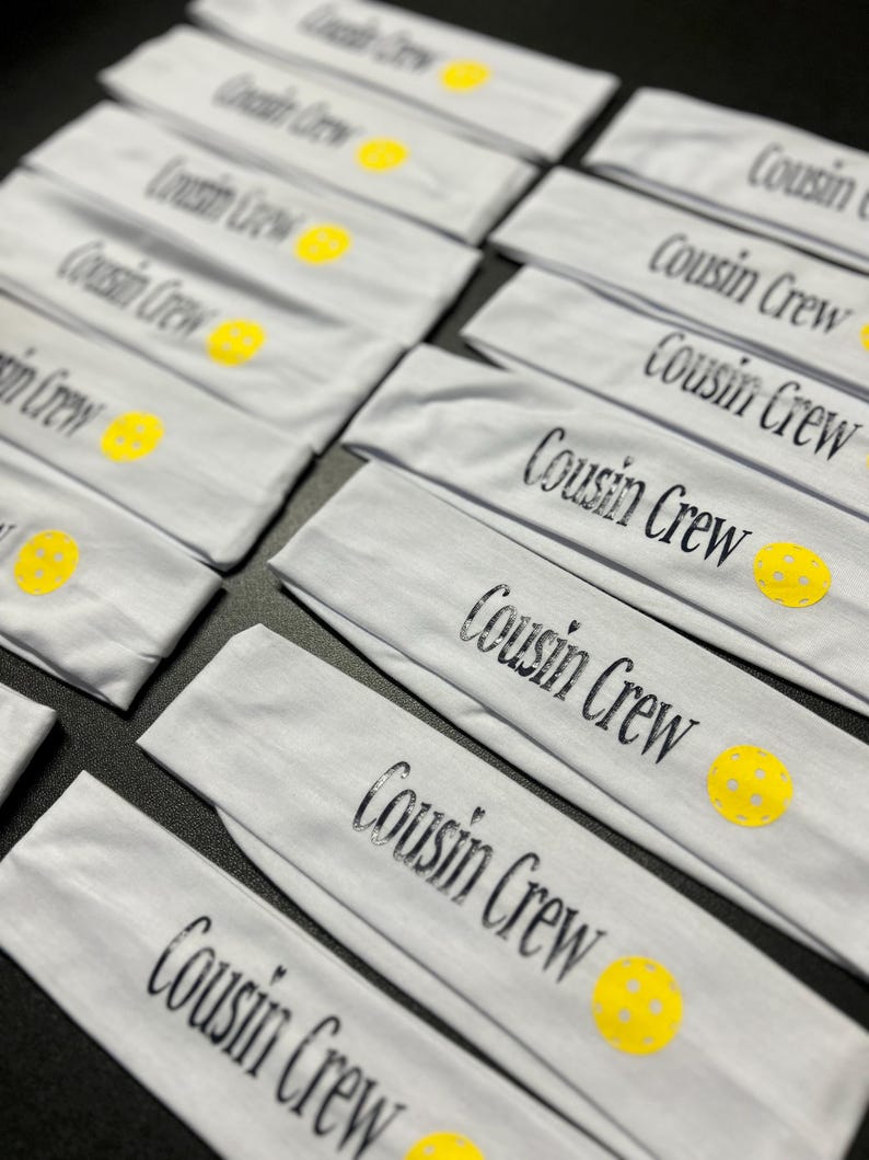 May include: A collection of light gray headbands with the text "Cousin Crew" in black script. Each headband features a yellow pickleball design. The headbands are neatly arranged, suggesting they are ready for sale or distribution.