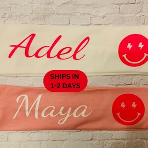 May include: Two headbands, one white and one peach, each with a name and a red smiley face with lightning bolts. The white headband says "Adel" in red script. The peach headband says "Maya" in white script.