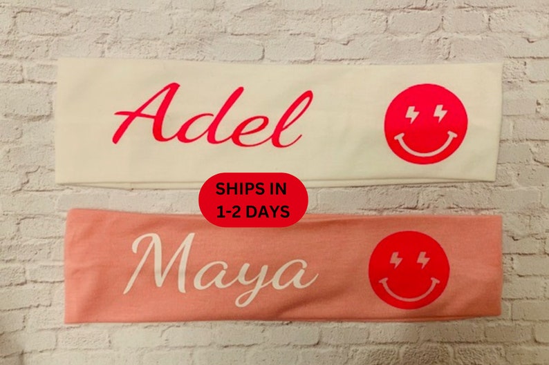 May include: Two headbands, one white and one peach, each with a name and a red smiley face with lightning bolts. The white headband says "Adel" in red script. The peach headband says "Maya" in white script.