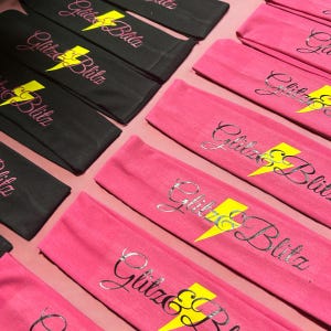 May include: Rows of black and pink headbands with the text "Glitz & Blitz" in a cursive font and a yellow lightning bolt graphic. The headbands are made of a soft material and are arranged neatly.