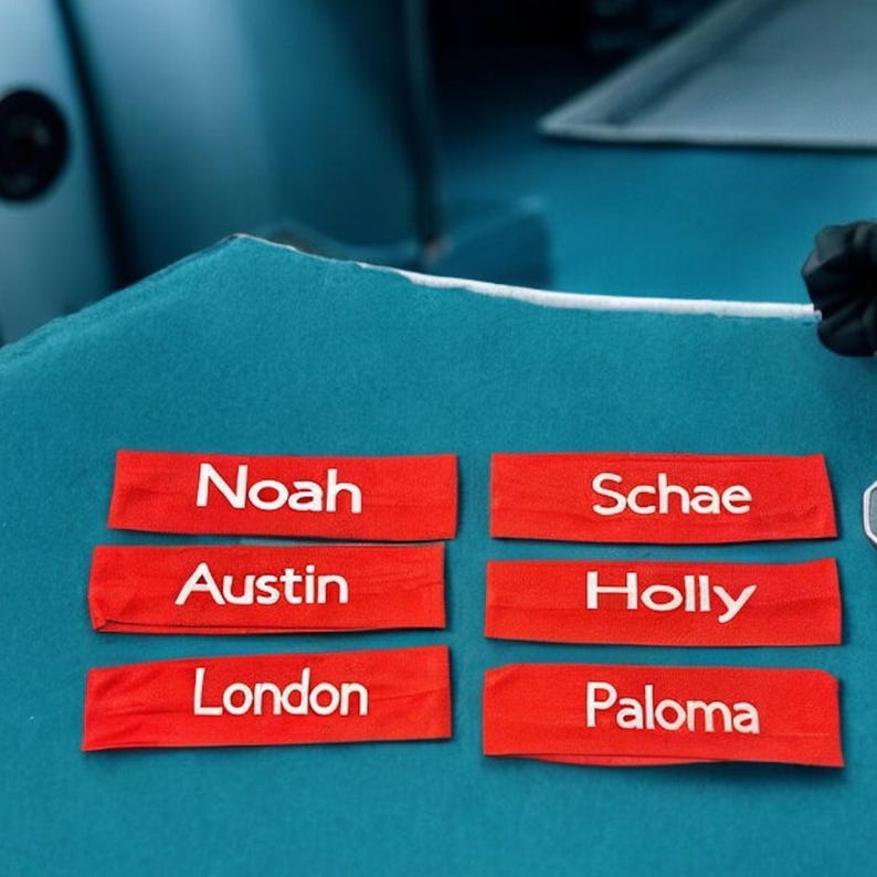 May include: Six red fabric name tags with white lettering are arranged on a teal surface. The names on the tags are Noah, Austin, London, Schae, Holly, and Paloma. The background is a solid teal color.