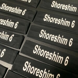 May include: Black headbands with the text "Shoreshim 6" printed in white. The headbands are rectangular and appear to be made of a soft, stretchy material. Multiple headbands are arranged closely together.