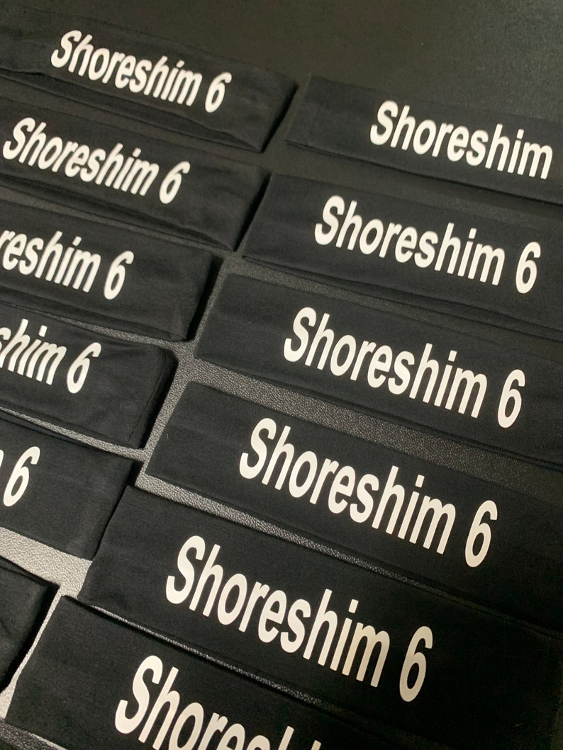 May include: Black headbands with the text "Shoreshim 6" printed in white. The headbands are rectangular and appear to be made of a soft, stretchy material. Multiple headbands are arranged closely together.