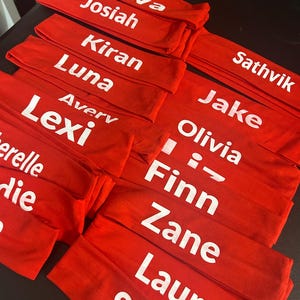 May include: Several red headbands with white text displaying various names. The names include Ava, Josiah, Kiran, Luna, Avery, Lexi, Sathvik, Jake, Olivia, Finn, Zane, Laura, and Silvi. The headbands are neatly stacked.