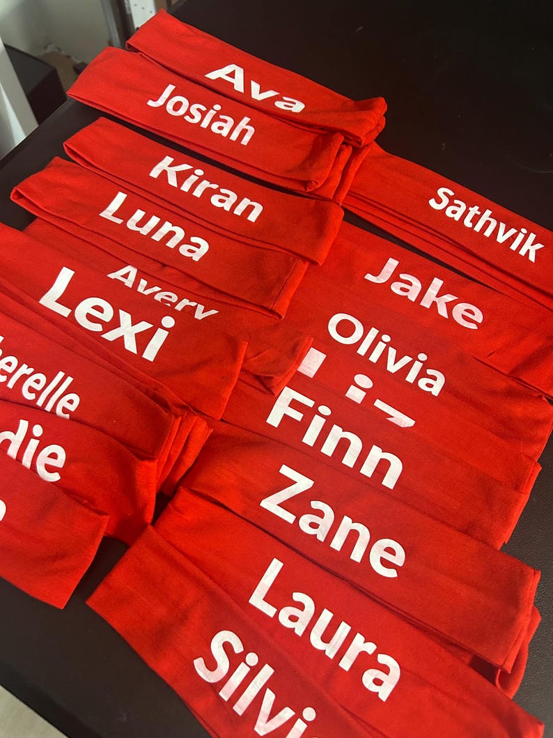 May include: Several red headbands with white text displaying various names. The names include Ava, Josiah, Kiran, Luna, Avery, Lexi, Sathvik, Jake, Olivia, Finn, Zane, Laura, and Silvi. The headbands are neatly stacked.