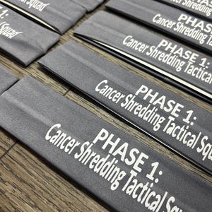 May include: Several gray headbands are arranged on a wooden surface. Each headband has the text "PHASE 1: Cancer Shredding Tactical Squad" printed in white. The headbands are folded and appear to be made of a soft material.