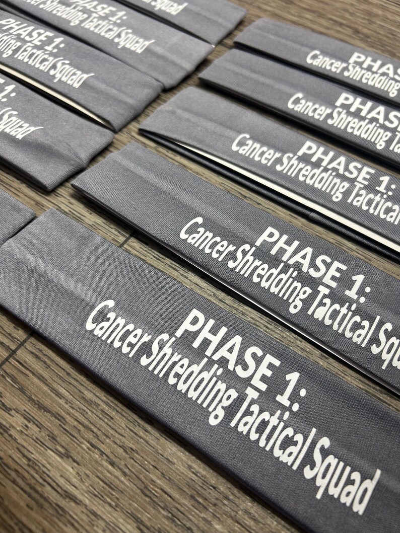 May include: Several gray headbands are arranged on a wooden surface. Each headband has the text "PHASE 1: Cancer Shredding Tactical Squad" printed in white. The headbands are folded and appear to be made of a soft material.