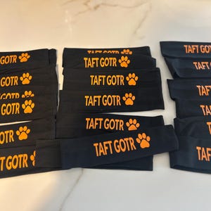 May include: Black headbands with the text "TAFT GOTR" in orange, along with an orange paw print. The headbands are stacked and arranged on a light-colored surface. The headbands are likely for athletic use.