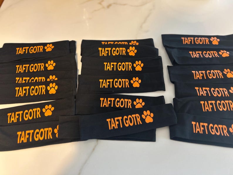 May include: Black headbands with the text "TAFT GOTR" in orange, along with an orange paw print. The headbands are stacked and arranged on a light-colored surface. The headbands are likely for athletic use.