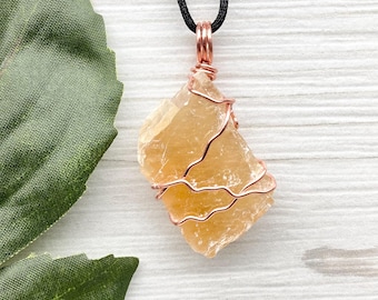 May include: A raw, light orange calcite crystal wrapped in copper wire, hanging from a black cord necklace. The crystal is a natural, irregular shape.