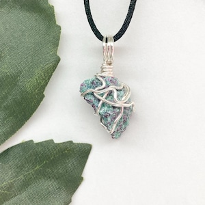 May include: A silver wire wrapped pendant featuring a green and purple gemstone. The pendant is hanging from a black cord.