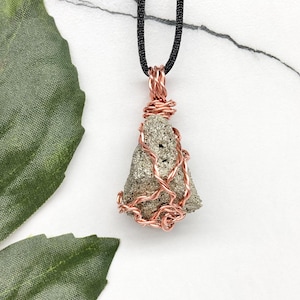 May include: A pyrite pendant with a copper wire wrap, suspended from a black cord. The pyrite stone has a metallic, speckled appearance. The copper wire is intricately woven around the stone, creating a unique, handcrafted necklace.