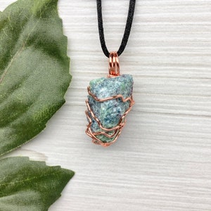 May include: A pendant necklace featuring a rough-cut, blue-green stone wrapped in copper wire. The stone hangs from a black cord. The copper wire is wrapped around the stone, creating a cage-like design. The background is a light wood grain.