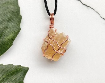 May include: A raw, golden calcite crystal pendant wrapped in copper wire, suspended from a black cord. The crystal has a rough, natural texture and is a warm, amber color. The pendant is a unique piece of jewelry.