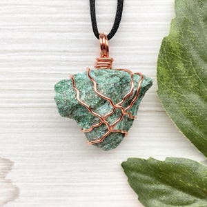 May include: A green stone pendant necklace. The stone is wrapped in copper wire and suspended from a black cord. The stone has a rough texture and is a mix of green hues. The copper wire adds a decorative element to the necklace.
