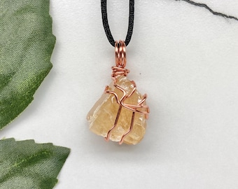 May include: A raw, golden-yellow citrine crystal pendant wrapped in copper wire, suspended from a black cord. The gemstone is irregularly shaped and has a natural, unpolished appearance. The pendant is a unique piece of jewelry.