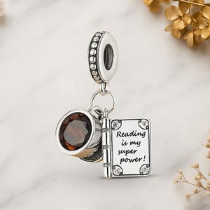 May include: A silver charm with a round, dark brown gemstone, a book charm with the text "Reading is my super power!", and a decorative bead. The charm is designed to be added to a bracelet or necklace.