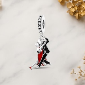 Könnte beinhalten: Silberner Charm mit einem Design eines tanzenden Paares. Der Charm hat einen schwarzen Smoking, ein rotes Kleid und einen silbernen Bügel, der mit klaren Kristallen verziert ist. Ein romantisches Accessoire.