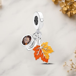 Può includere: Un charm in argento con una tazza di caffè e una foglia autunnale. La tazza di caffè ha una gemma marrone e la parola "coffee". La foglia è arancione e gialla.